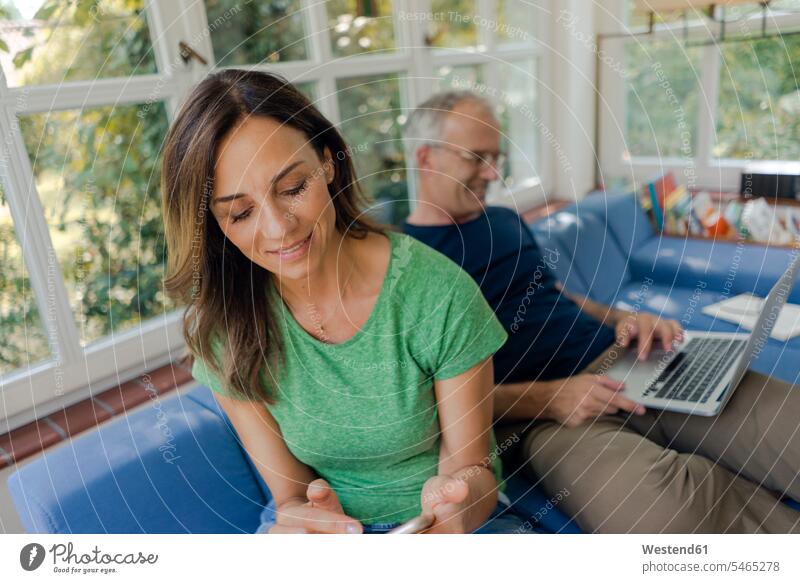 Mature couple sitting on couch at home with woman using cell phone and man using laptop couches settee settees sofa sofas Seated computers Laptop Computer