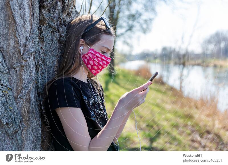 Girl with homemade protective mask using smartphone and leaning on tree telecommunication phones telephone telephones cell phone cell phones Cellphone mobile