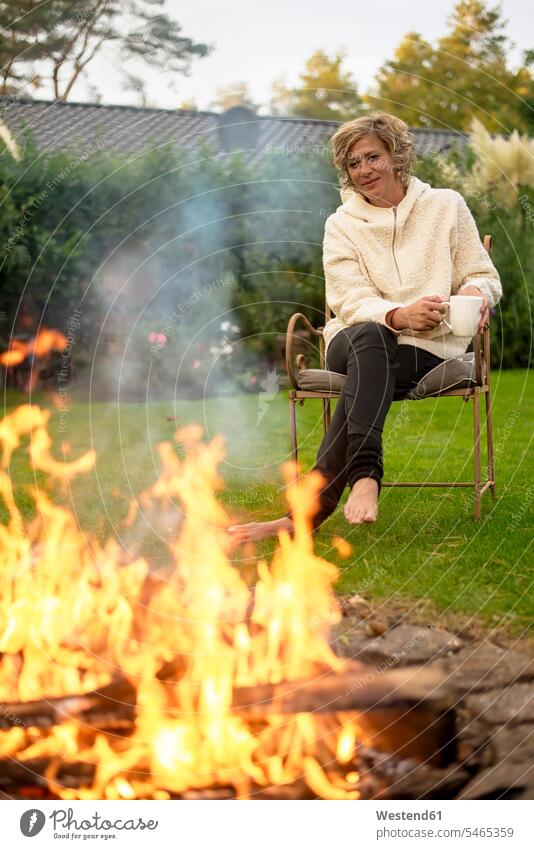 Mature woman with coffee cup looking at bonfire while sitting on chair at back yard color image colour image outdoors location shots outdoor shot outdoor shots