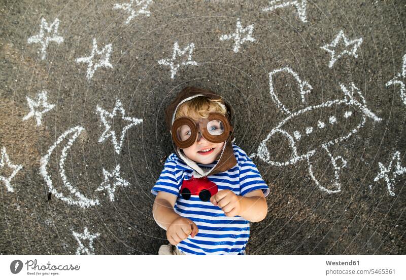 Portrait of smiling toddler wearing pilot hat and goggles lying on asphalt painted with airplane, moon and stars aeroplanes airplanes the moon moons portrait