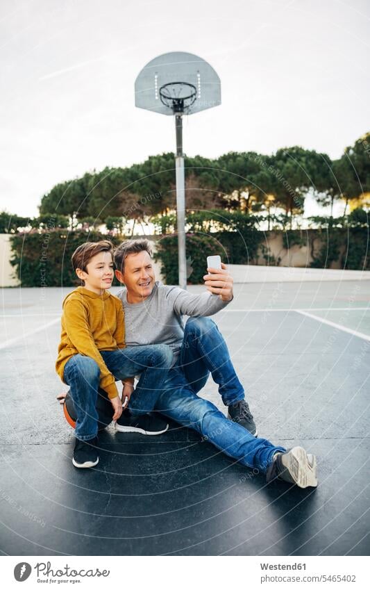 Father and son sitting on basketball outdoor court taking a selfie sports field sports fields Seated father pa fathers daddy dads papa Selfie Selfies sons