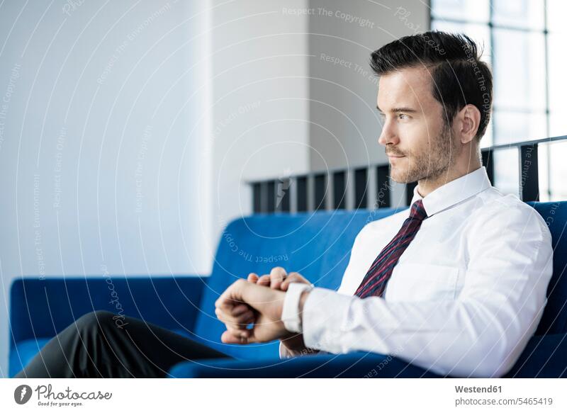 Businessman sitting on blue couch in office checking smartwatch Seated Business man Businessmen Business men offices office room office rooms smart watch