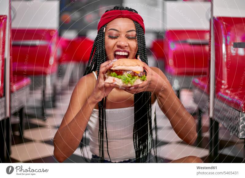 Young woman with braided hairstyle sitting on the floor and grabbing a big hamburger with her two hands Seated Sitting On Floor Sitting On The Floor land smile