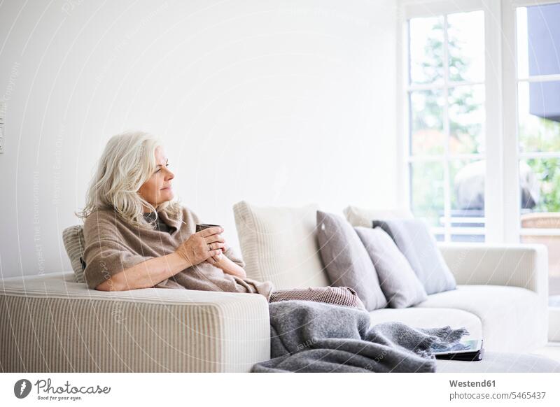 Thoughtful woman with long white hair holding coffee cup while sitting on sofa at apartment color image colour image Denmark Scandinavia Scandinavian Peninsula