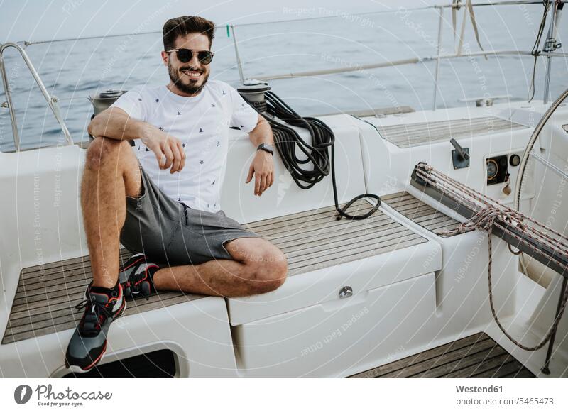 Smiling young man wearing sunglasses sitting in sailboat color image colour image Spain outdoors location shots outdoor shot outdoor shots day daylight shot