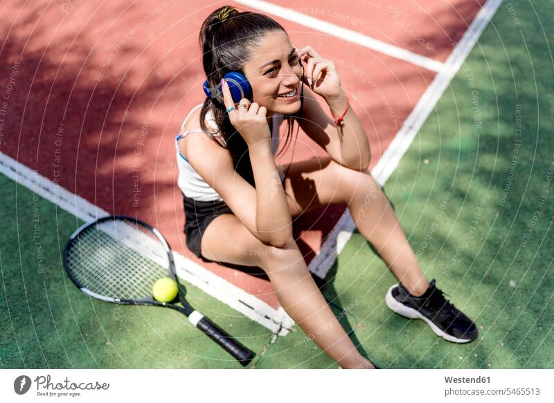 Smiling female tennis player listening music while sitting on floor in court color image colour image Spain outdoors location shots outdoor shot outdoor shots