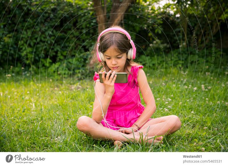 Portrait of girl sitting on a meadow using smartphone and pink headphones magenta females girls headset Seated Smartphone iPhone Smartphones use meadows colour