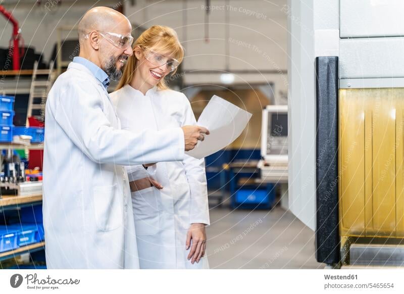 Two happy technicians wearing lab coats and safety glasses looking at plan in factory plans factories Protective Eyeglasses safety goggles Protective Glasses