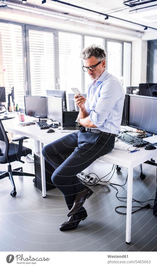 Mature businessman sitting on desk in office using smartphone Seated offices office room office rooms Smartphone iPhone Smartphones desks Businessman