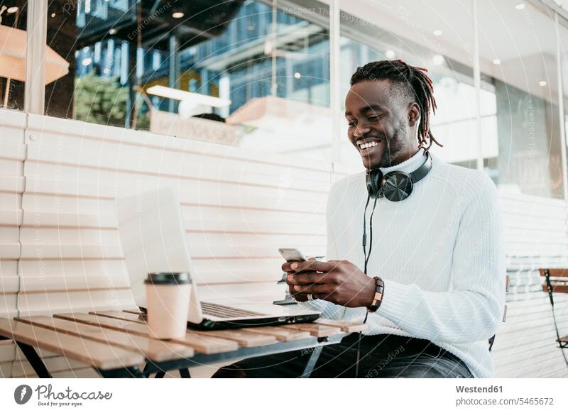 Smiling creative businessman with laptop using mobile phone in cafe color image colour image outdoors location shots outdoor shot outdoor shots day