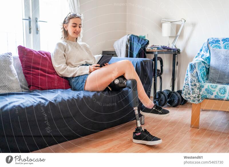 Young woman with leg prosthesis sitting on couch at home reading e-book human human being human beings humans person persons celibate celibates singles