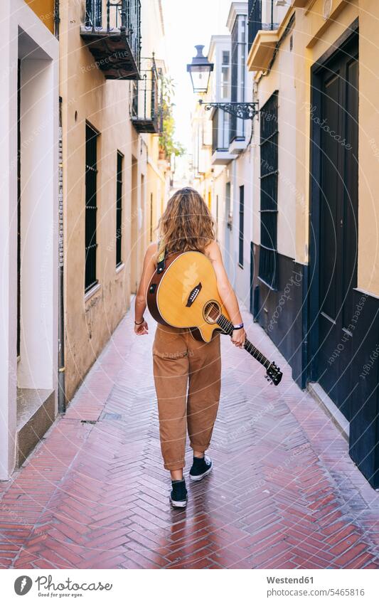 Rear view of young woman with guitar walking on narrow street amidst buildings at Santa Cruz, Seville, Spain color image colour image Sevilla Andalusia