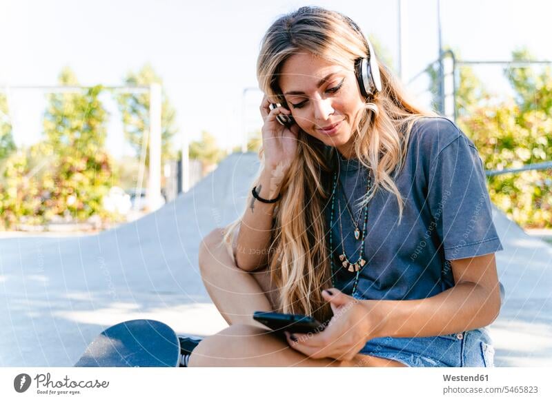 Young blond woman listening music through headphones while using smart phone at skateboard park color image colour image outdoors location shots outdoor shot