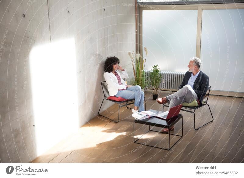 Businessman and businesswoman sitting in a loft at concrete wall talking speaking Business man Businessmen Business men Seated concrete walls businesswomen