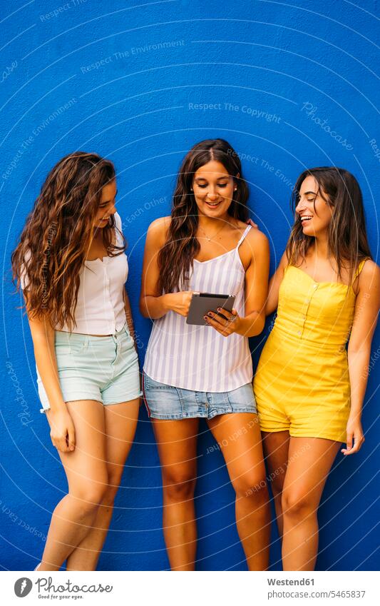 Three happy female friends standing at a blue wall sharing tablet mate dark-haired brown haired brunette video hold smile speak speaking talk seasons