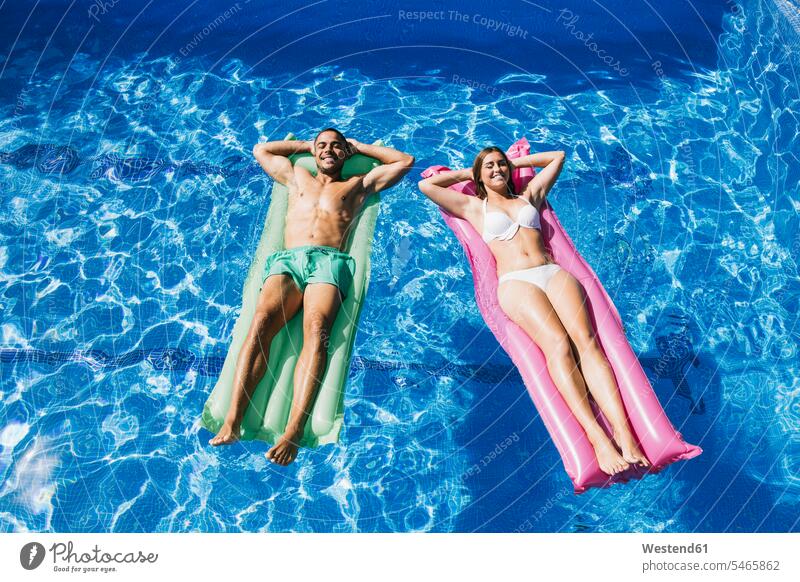 Smiling young couple with hands behind head relaxing on airbeds in swimming pool color image colour image Spain day daylight shot daylight shots day shots