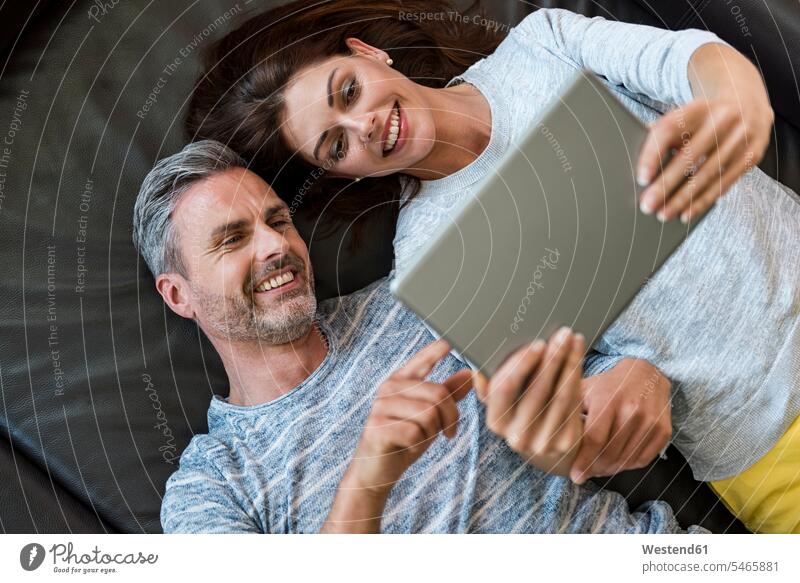 Happy couple lying on couch at home sharing a tablet share twosomes partnership couples happiness happy settee sofa sofas couches settees digitizer