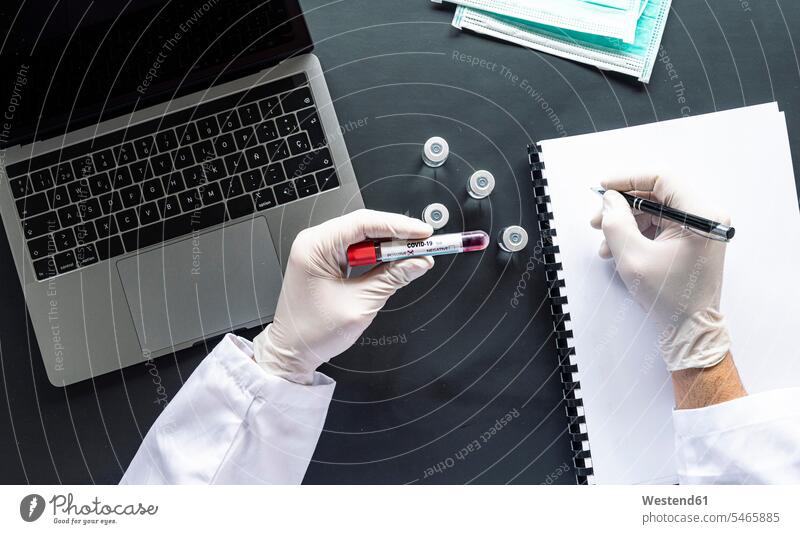 Doctor with COVID-19 sample writing on spiral notebook by laptop at desk in laboratory color image colour image indoors indoor shot indoor shots interior