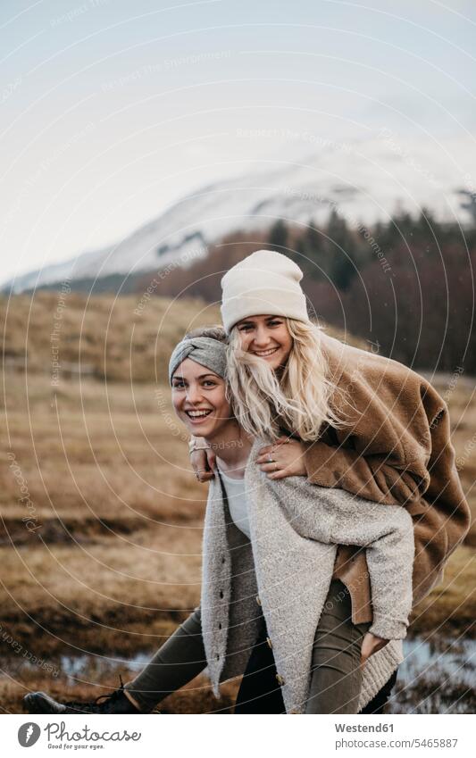 UK, Scotland, happy young woman carrying friend piggyback in rural landscape female friends portrait portraits piggy-back pickaback Piggybacking Piggy Back