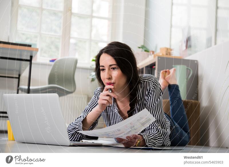 Casual woman with sheet of paper and laptop lying on the floor in office females women offices office room office rooms laying down lie lying down floors sheets