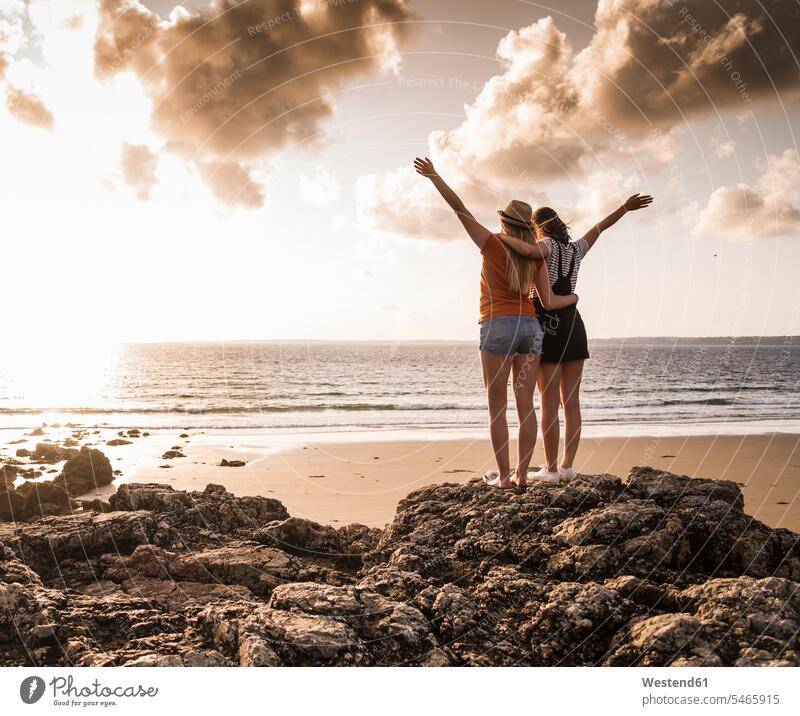 Two girlfriends standing on rocky beach, waving at sunset, rear view delight enjoyment Pleasant pleasure Cheerfulness exhilaration gaiety gay glad Joyous merry