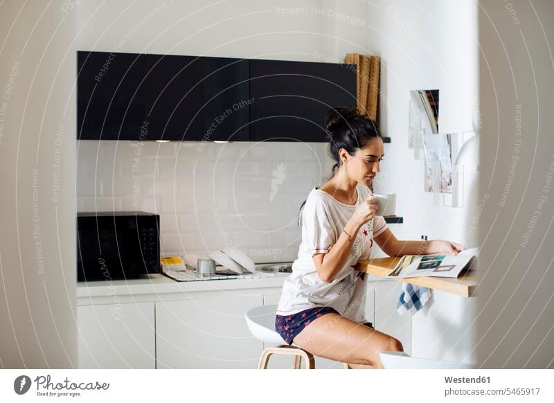 Young woman reading a magazine and drinking coffee in kitchen human human being human beings humans person persons celibate celibates singles solitary people