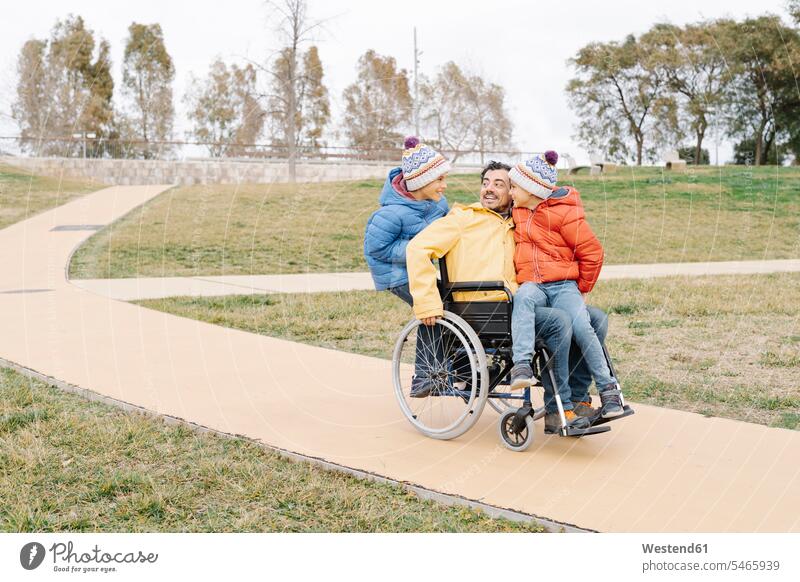 Playful man with excited sons riding wheelchair on footpath in park color image colour image Spain leisure activity leisure activities free time leisure time