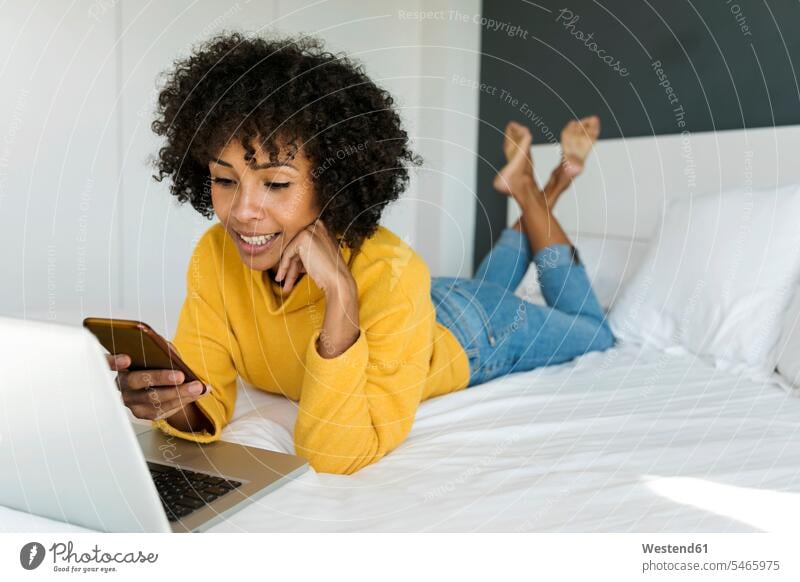 Smiling woman lying on bed using cell phone and laptop Laptop Computers laptops notebook mobile phone mobiles mobile phones Cellphone cell phones laying down