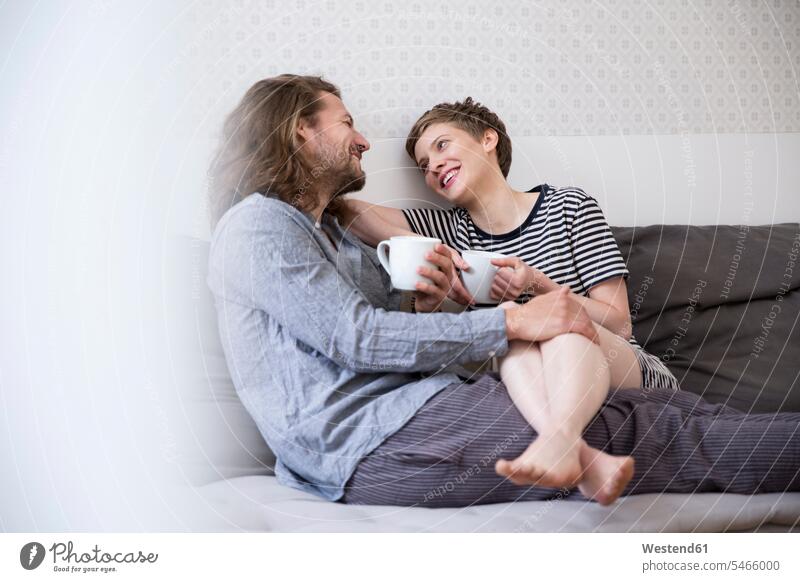 Happy young couple in pyjamas on bed couch at home couches settee settees sofa sofas hold cuddle snuggle snuggling smile Seated sit drink in the morning relax