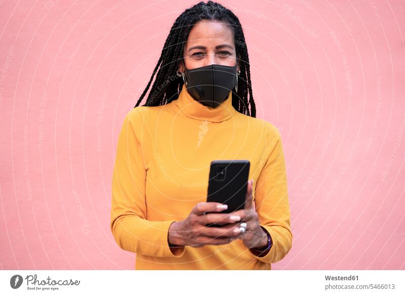 Mature woman wearing face mask using mobile phone while standing against pink wall color image colour image outdoors location shots outdoor shot outdoor shots