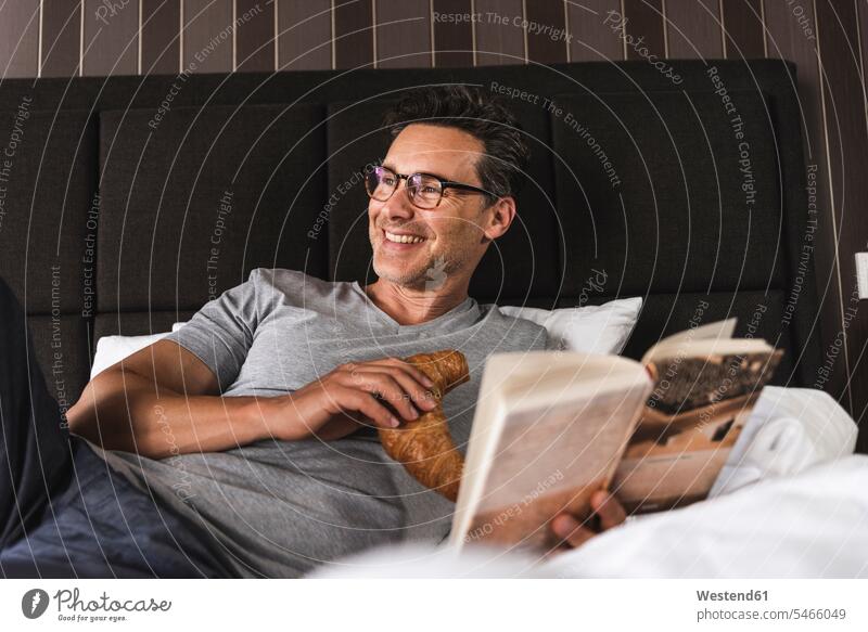 Happy man lying in bed at home with croissant and book happiness happy men males beds laying down lie lying down reading books Croissant Croissants Cornetto