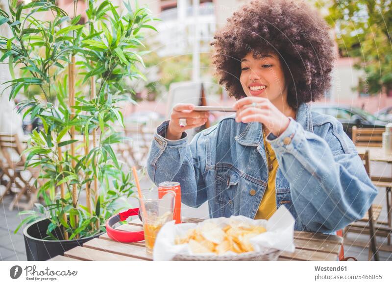 Smiling young woman with afro hairdo taking smartphone picture at an outdoor cafe in the city human human being human beings humans person persons curl curled