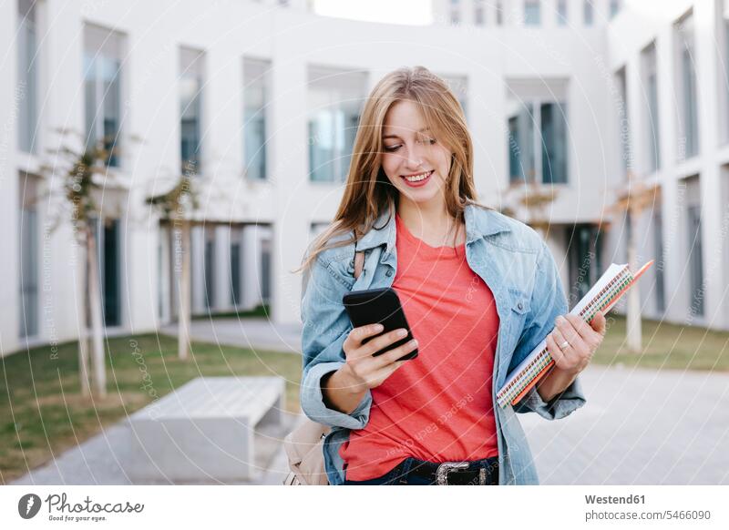 Smiling young blond female student using smart phone at university beautiful Woman beautiful Women people human being human beings humans person persons Campus