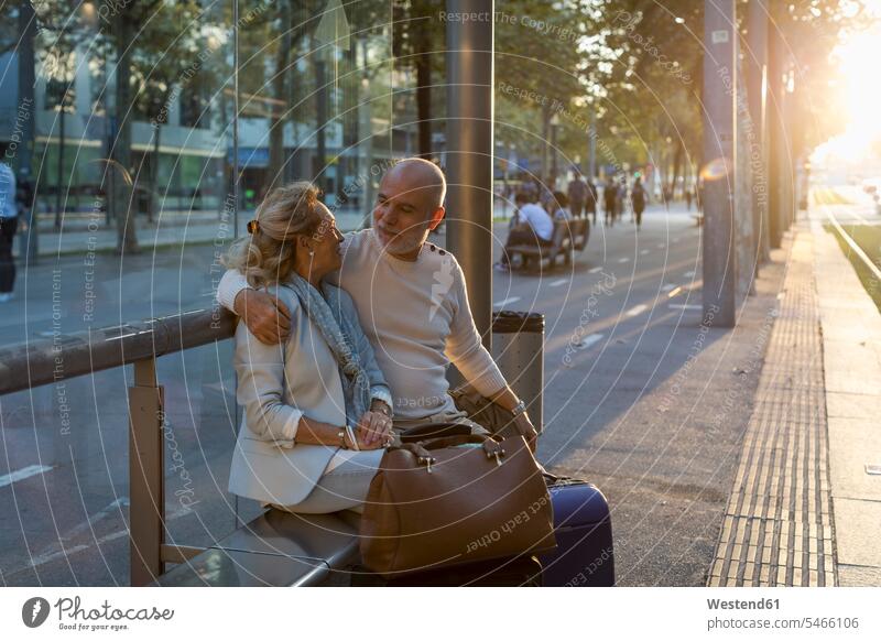 Spain, Barcelona, senior couple with baggage sitting at tram stop in the city at sunset tram station tram stops tram stations town cities towns sunsets sundown