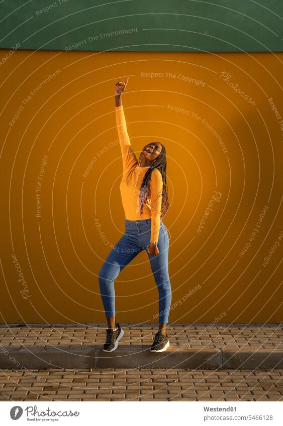 Teenage girl dancing with hand raised against yellow wall on street color image colour image outdoors location shots outdoor shot outdoor shots day