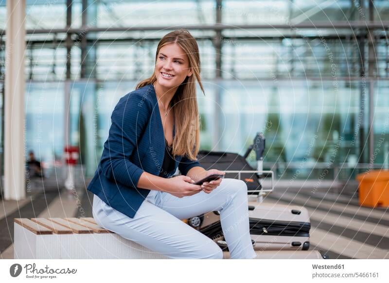 Smiling young businesswoman sitting outdoors with cell phone and suitcase looking around mobile phone mobiles mobile phones Cellphone cell phones Seated