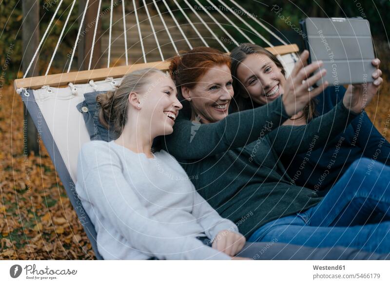 Happy mother with two teenage girls lying in hammock in garden in autumn using tablet fall laying down lie lying down hammocks mommy mothers ma mummy mama