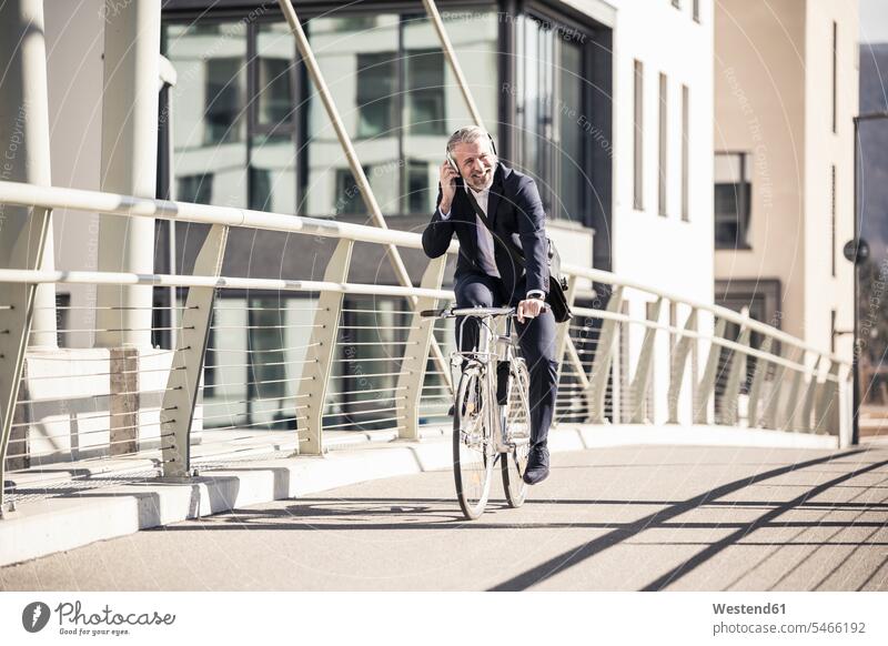 Smiling mature businessman with headphones riding bicycle on a bridge in the city bridges town cities towns smiling smile headset bikes bicycles Businessman
