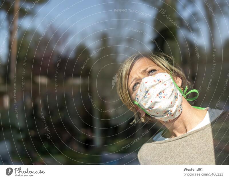 Woman with face mask looking through window glass on sunny day during quarantine color image colour image domestic life at home domestic scene in quarantine