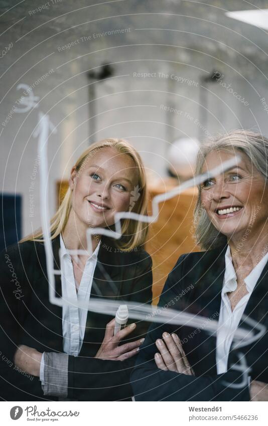 Happy mature and young businesswoman looking at chart on glass pane in office human human being human beings humans person persons caucasian appearance
