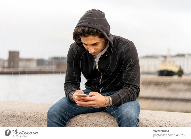 Denmark, Copenhagen, young man sitting on wall at the waterfront using cell phone Kobenhavn chatting Smartphone iPhone Smartphones Traveller Travellers