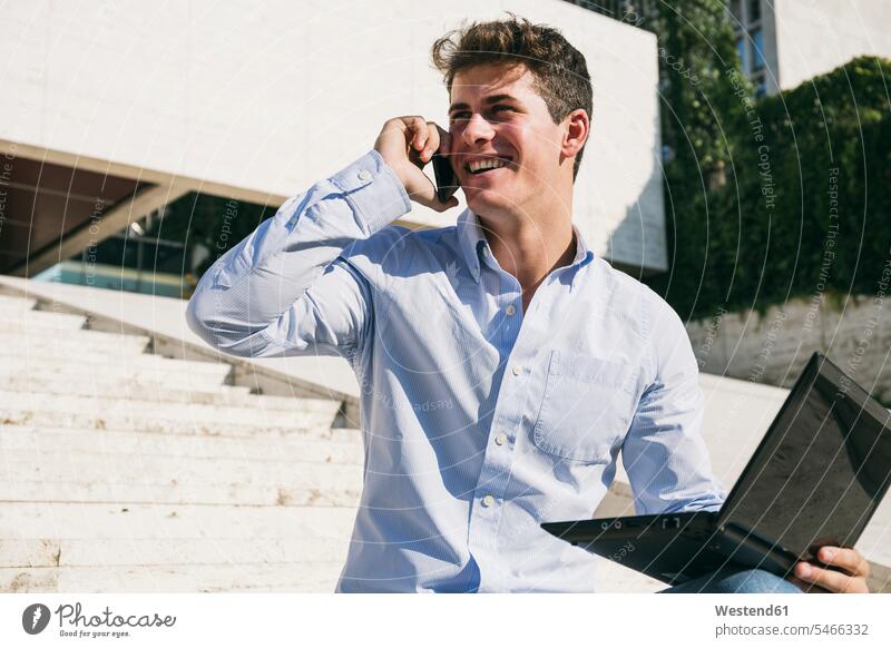 Smiling young man talking on mobile phone while sitting with laptop during sunny day color image colour image Spain outdoors location shots outdoor shot