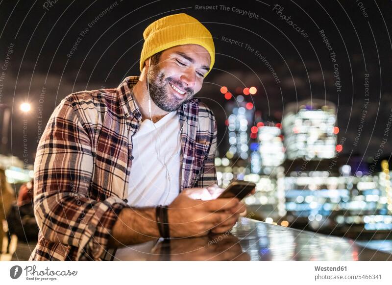 UK, London, smiling man leaning on a railing and looking at his phone with city lights in background men males smile night by night at night nite
