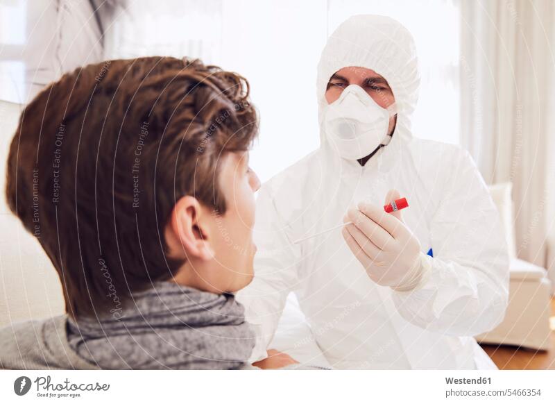 Doctor wearing protective suit doing Coronavirus test of young man at home color image colour image Germany indoors indoor shot indoor shots interior