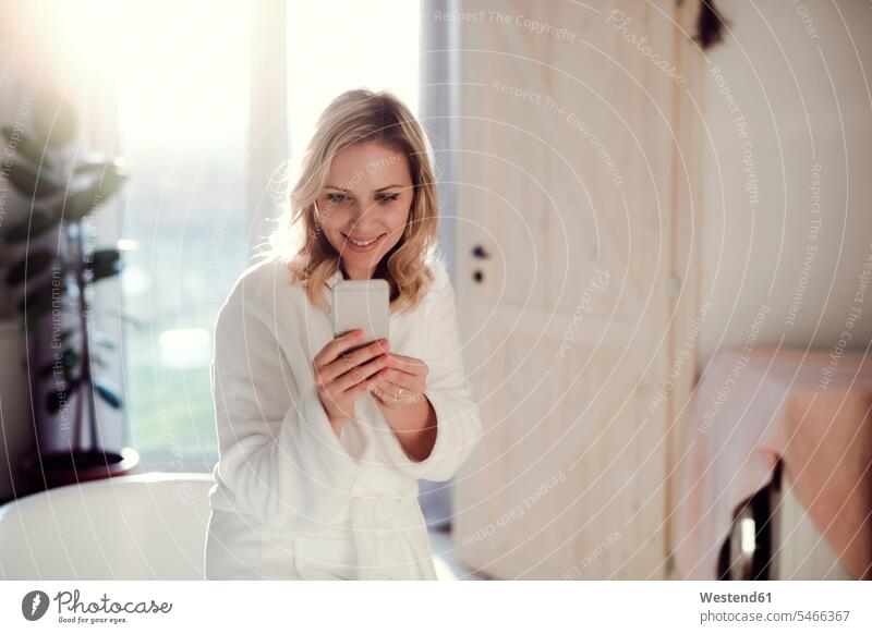 Smiling woman wearing bathrobe in bathroom at home holding cell phone females women Domestic Bathroom bath room morning in the morning smiling smile