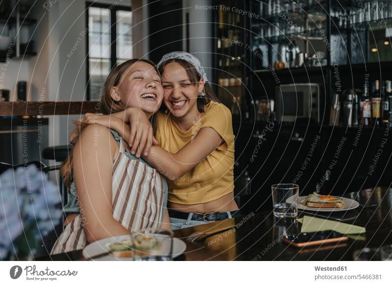 Teenage girlfriends sitting at dining table, embracing and laughing Glasses dish dishes Plates Tables Dining Tables Dinner Table smile Seated embrace
