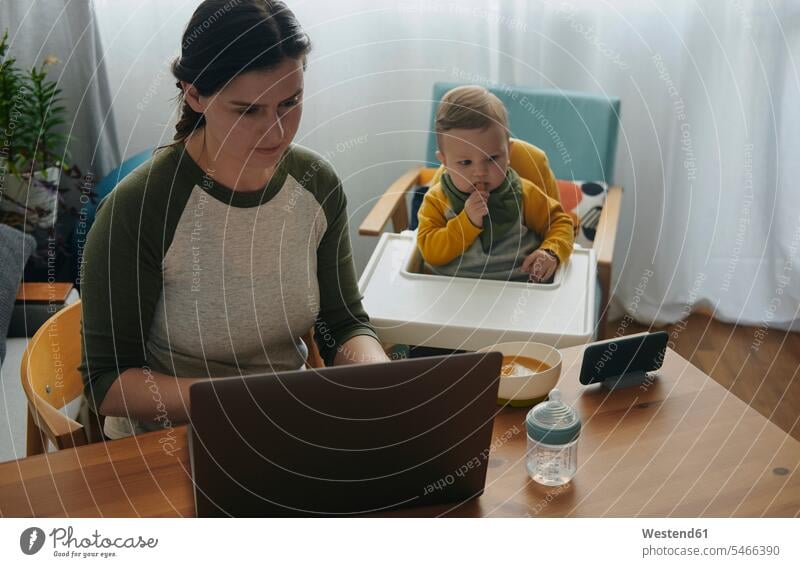 Woman working on laptop at home while her little son watching movies on mobile phone human human being human beings humans person persons caucasian appearance