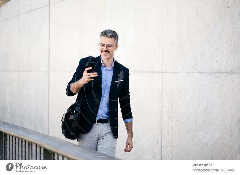 Smiling gray-haired businessman walking in the city using cell phone business life business world business person businesspeople Business man Business men