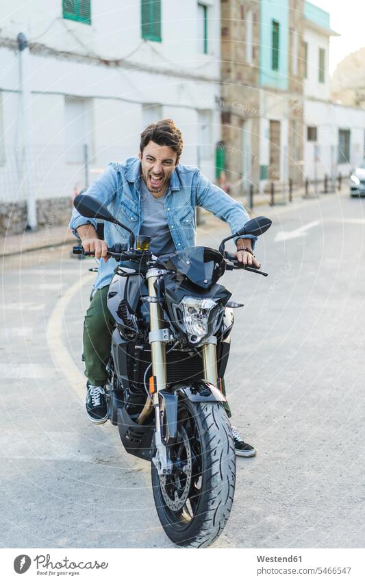 Portrait of screaming young man sitting on motorbike pulling funny faces shouting grimace grimacing grimaces Motor Cycle men males portrait portraits Seated