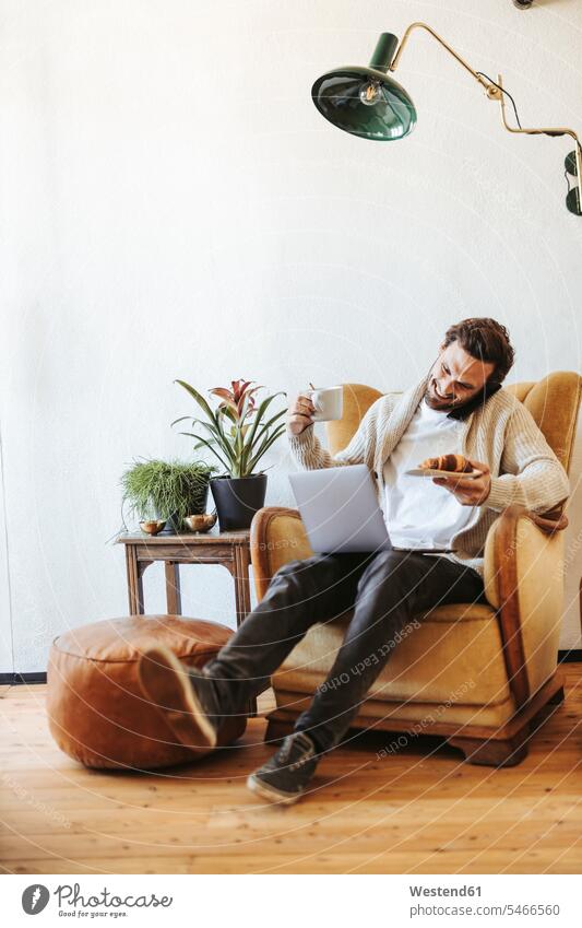 Smiling man on the phone sitting on armchair at home with laptop, croissant and cup of coffee human human being human beings humans person persons